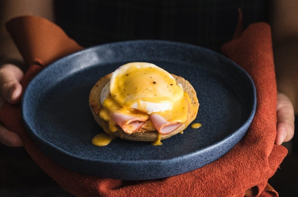egg Benedict with hollandaise sauce on dark blue plate on table in kitchen